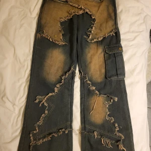 Unika slitna jeans med fransar - Säljer ett par riktigt coola jeans med en unik design. De är mörkblå med slitna detaljer och fransar över hela byxorna. Det finns också en praktisk cargoficka på sidan. Perfekta för dig som vill sticka ut med en edgy stil!