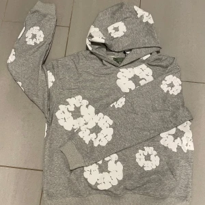 Grå hoodie från Denim Tears - Säljer en supercool grå hoodie från Denim Tears med vita blommönster över hela tröjan. Den är i nytt skick och perfekt för både höst och vår. Hoodien har en klassisk passform med långa ärmar och en mysig huva. Perfekt för att hålla stilen när det är lite kyligare ute! 🌸