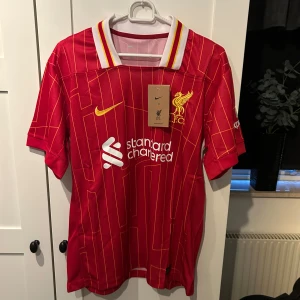 Röd fotbollströja från Nike - Säljer en röd fotbollströja från Nike med gula och vita detaljer. Tröjan har korta ärmar och ett randigt mönster. Den är i nytt skick och har en klassisk krage. Perfekt för fotbollsfans som vill visa sitt stöd!