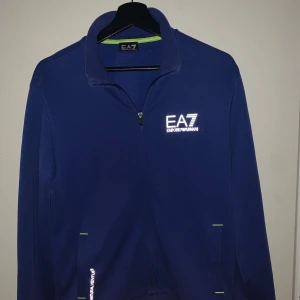 Blå tröja från Emporio Armani EA7 - Säljer en snygg blå tröja från Emporio Armani EA7. Den har en dragkedja framtill och ett reflekterande tryck på bröstet och ryggen. Perfekt för en sportig look eller en avslappnad dag. Tröjan är i mycket bra skick och har en normal passform. Äkta med qr-kod som verifierar äkthet!