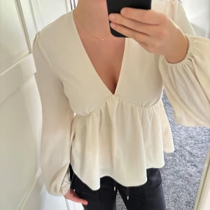 Beige blus från NA-KD - Säljer en superfin beige blus från NA-KD i mycket bra skick. Den har en v-ringad design och långa ärmar med en lätt volangdetalj i midjan. Perfekt för både vardag och festliga tillfällen. Passar bra till både jeans och kjol!
