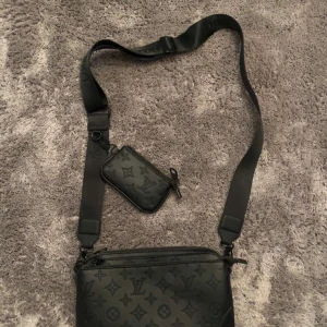 Svart axelväska med monogram - Snygg svart axelväska med monogrammönster. Väskan har en justerbar axelrem och en mindre avtagbar väska. Perfekt för både vardag och fest. Den är i mycket bra skick och har en dragkedja för säker förvaring.