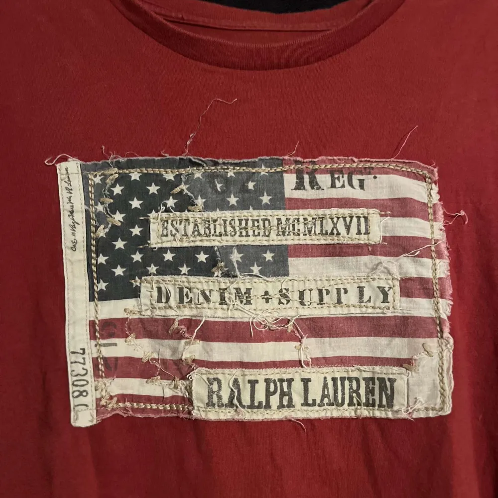 Säljer en röd t-shirt från Ralph Lauren med ett vintageinspirerat tryck av den amerikanska flaggan och texten 'Denim & Supply'. T-shirten är i 100% bomull och har en avslappnad passform. Perfekt för en casual look!. T-shirts.