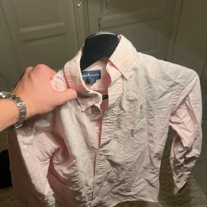 Rosa och vit randig skjorta från Ralph Lauren - Säljer en snygg rosa och vit randig skjorta från Ralph Lauren. Skjortan är långärmad och har klassiska knappar. Den är i 10/10 skick och gjord av bomull. Skjortan är i stl 12 (xs). Pris kan diskuteras vid snabb affär.