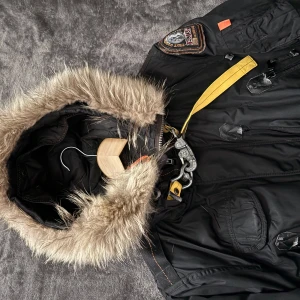 Svart vinterjacka med pälsdetaljer - Säljer en svart vinterjacka från Parajumpers med pälsfodrad huva. Jackan har flera fickor och en cool gul remdetalj vid halsen. Perfekt för kalla dagar! 🖤Butikskvutto finns köptes för 8900kr 