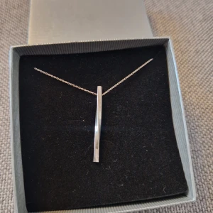 Silverhalsband med minimalistisk hängsmycke - Säljer ett elegant silverhalsband med ett stilrent, avlångt hängsmycke. Halsbandet är i 925 sterling silver och har en tunn kedja som ger en minimalistisk look. Perfekt för både vardag och fest. Kommer i en fin presentask.