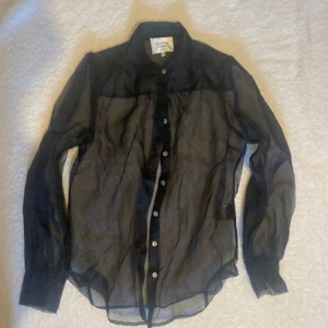 Svart blus från Second Female - Snygg svart transparent blus från Second Female i storlek XS. Perfekt för en festlig kväll eller som ett lager över ett linne. Blusen har långa ärmar och knappar framtill. Den är både stilren och trendig, vilket gör den till ett måste i garderoben. Använd få gånger!