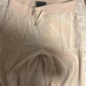 Beige mjukisbyxor från Juicy Couture - Säljer ett par superbekväma beige mjukisbyxor från Juicy Couture i storlek M. Byxorna har en hög midja med snörning och två fickor fram och bak med knappar. Perfekta för en chill dag hemma eller en avslappnad utflykt. Broderad logga på ena benet för en extra touch av stil. Nästan aldrig använd💓