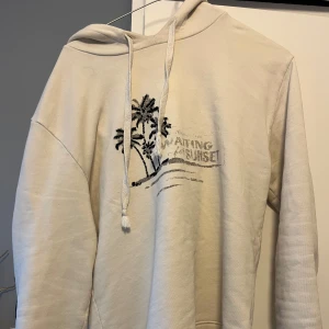 Beige hoodie från Saint Laurent - Säljer en snygg beige hoodie från Saint Laurent med tryck av palmer och texten 'Waiting for Sunset'. Den är supermjuk och perfekt för en chill dag. Hoodien har en klassisk passform med dragsko i huvan. Perfekt för både höst och vår!