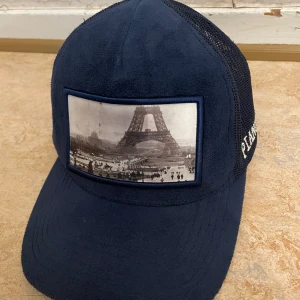 Blå keps med Eiffeltornet från Pianotti - Snygg blå keps från Pianotti med ett coolt tryck av Eiffeltornet på framsidan. Kepsen har ett nätmaterial på baksidan för bra ventilation och märkesnamnet broderat på sidan. Perfekt för en avslappnad stil eller en dag ute i solen! Priset kan prutas till en viss nivå.  Har inte använt kepsen mycket och vill sälja den för att jag inte behöver den längre.