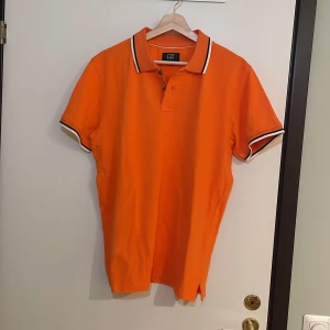 Orange pikétröja från Cutter & Buck - Säljer en snygg orange pikétröja från Cutter & Buck i storlek L. Tröjan har en klassisk krage med knappar och vita detaljer på krage och ärmslut. Perfekt för sommardagar eller en avslappnad dag på stan. 🧡 Pris går alltid att förhandlas!