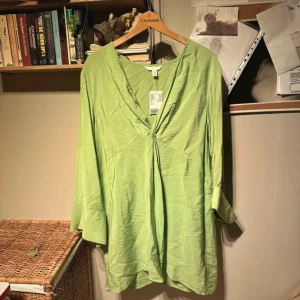 Grön blus från H&M - Säljer en fräsch grön klänning från H&M i storlek L. Blusen har en v-ringad design och är tillverkad i ett lätt och luftigt material, perfekt för vår och sommar. Den har en lös passform och trekvartsärmar, vilket gör den både bekväm och stilren.