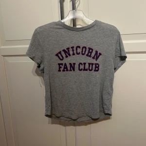 Grå t-shirt med 'Unicorn Fan Club' tryck - Säljer en grå t-shirt i bomull med texten 'Unicorn Fan Club' i blå och röd på framsidan. Den är kortärmad och har en croppad passform, perfekt för en avslappnad stil. T-shirten är i bra skick och passar perfekt till vardags.