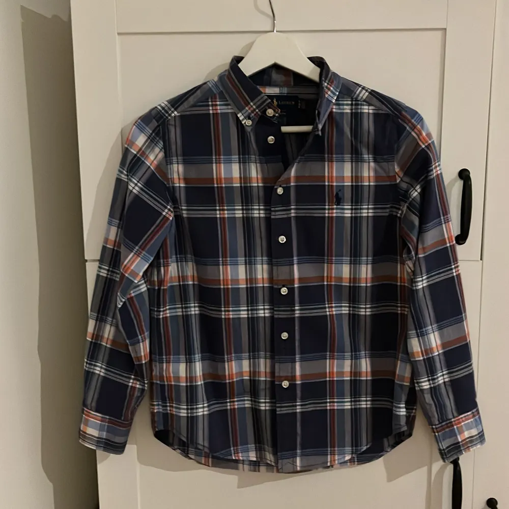 Säljer en snygg rutig skjorta från Ralph Lauren som passar barn 10-12 år. Skjortan har ett klassiskt rutmönster i blått, orange, vitt och grönt. Den är långärmad och har knappar framtill. Perfekt för både vardag och fest! 🕺. Skjortor.