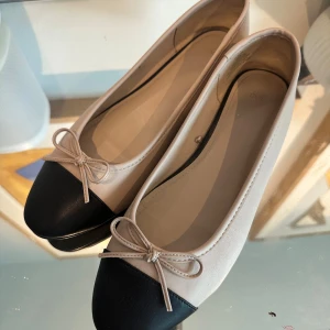 Beige och svarta ballerinaskor med rosett - Säljer ett par eleganta ballerinaskor i beige med svart tå och söt rosett. Perfekta för både vardag och fest! De är i mycket bra skick och har en klassisk design som passar till allt. Skorna har en bekväm passform och är lätta att bära hela dagen.