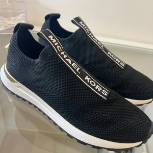 Svarta sneakers från Michael Kors - Snygga svarta sneakers från Michael Kors i stickat material med vit sula och guldiga detaljer. De har en elastisk passform och tydlig logga på ovansidan. Perfekta för en stilren och bekväm look!