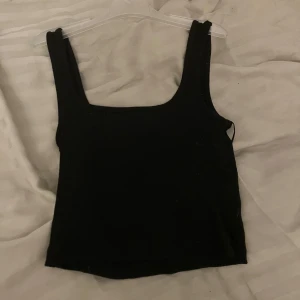 Svart ärmlös topp - Snygg svart ärmlös topp i ett enkelt och stilrent snitt. Perfekt för sommardagar eller som basplagg under en jacka. Toppen är croppad och har en tight passform som framhäver figuren. Ett måste i garderoben för en minimalistisk look!