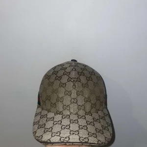 Beige keps med monogram - Snygg beige keps från Gucci med det klassiska monogrammönstret. Kepsen har en justerbar rem baktill i brunt läder och är perfekt för en stilren look.