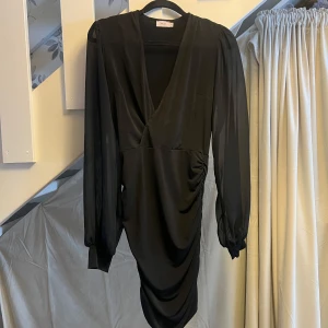 Svart omlottklänning från Nelly - Elegant svart omlottklänning från Nelly med långa ärmar och v-ringad design. Klänningen har en smickrande drapering framtill som ger en stilfull look. Perfekt för speciella tillfällen.