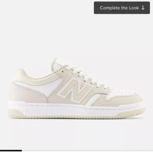 Beige och vita sneakers från New Balance - Snygga och stilrena sneakers från New Balance i beige och vitt. Skorna har en klassisk design med snörning och den ikoniska N-loggan på sidan. Perfekta för en avslappnad och trendig look.