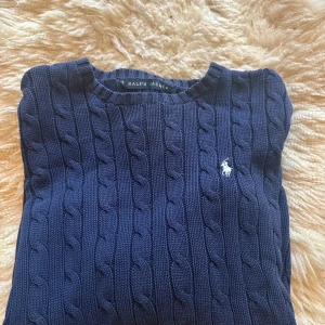 Mörkblå stickad tröja från Ralph Lauren - Säljer en snygg mörkblå stickad tröja från Ralph Lauren med kabelstickat mönster. Tröjan har långa ärmar och en klassisk rund halsringning. Perfekt för kyligare dagar!