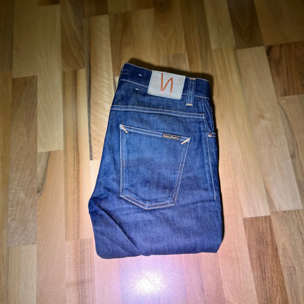 Säljer ett par mörkblå jeans från Nudie Jeans med klassisk femficksdesign och orange sömmar. Perfekta för en stilren look. Model: grim tim, storlek 31/32, finns små fläckar som ej syns på en distans. Farkut & Housut.
