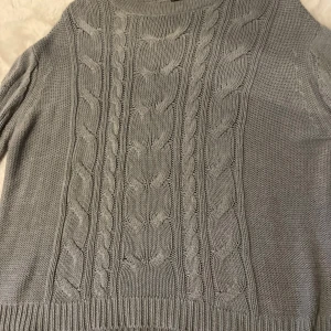 Grå stickad tröja från 157 - Mysig grå stickad tröja från 157 med kabelstickat mönster. Perfekt för kyliga dagar med sin klassiska design och långa ärmar. Ribbstickade muddar vid ärmslut och nederkant ger en skön passform. Grå blå färg 