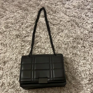 Svart quiltad axelväska från Pull&Bear - Snygg svart axelväska från Pull&Bear med quiltad design och kedjeaxelrem. Perfekt för att ge en stilren touch till din outfit. Väskan har en praktisk storlek och en elegant stängning framtill. Den är lite sliten på magnetstängningern💗
