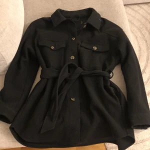 Svart overshirt med bälte - Snygg svart overshirt med knappar och knytbälte i midjan. Den har långa ärmar och två bröstfickor med knappar. Perfekt för lager-på-lager-stil.