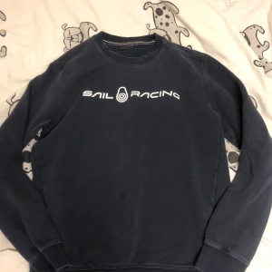 Mörkblå tröja från Sail Racing - Säljer en mörkblå tröja från Sail Racing med vit logga på bröstet. Tröjan har långa ärmar och rund halsringning. Perfekt för en avslappnad stil.