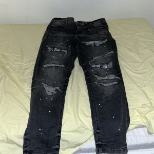Svarta jeans från Supply & Demand - Säljer ett par svarta jeans från Supply & Demand med slitna detaljer och färgstänk. De har en modern look med flera revor och en knappgylf. Perfekta för en edgy stil.