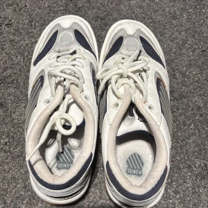 Vita sneakers från K-Swiss - Snygga vita sneakers från K-Swiss med marinblå och grå detaljer. Skorna har en klassisk snörning och en gummisula i orange. Perfekta för en sportig look. Squash utövare eller ej ☺️