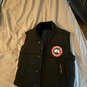 Svart dunväst från Canada Goose - Säljer en svart dunväst från Canada Goose med en ikonisk logga på bröstet. Västen har knappar och dragkedja framtill samt praktiska fickor. Perfekt för kyliga dagar.