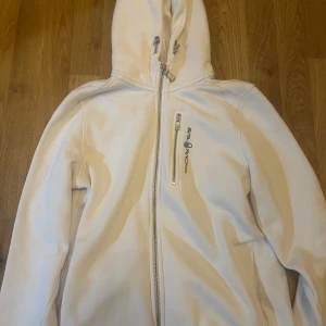 Vit fleecejacka från Sail Racing - Säljer en vit fleecejacka från Sail Racing med dragkedja och huva. Jackan har en broderad logga på bröstet och en praktisk bröstficka med dragkedja. Perfekt för kyliga dagar. Passar S