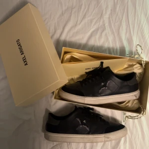 Svarta sneakers från Axel Arigato - Snygga svarta sneakers från Axel Arigato med vit sula och snörning (strl 45). Skorna har en stilren design med en fågel-logga på sidan. Perfekta för en trendig look. Små defekter på baksidan av skon. Rekommenderar att rengöra skorna också. Tveka inte på att höra av dig om du har någon fråga! 😉