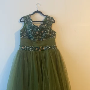 Grön långklänning med pärlor - Elegant grön långklänning med ett blommigt mönster på överdelen och pärldetaljer. Klänningen har en vid kjol och är ärmlös, perfekt för speciella tillfällen. Storleken kan ändras. 