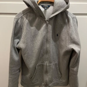 Grå hoodie från Ralph Lauren - Säljer en klassisk grå hoodie från Ralph Lauren med dragkedja och huva. Tröjan har en liten broderad logotyp på bröstet och är tillverkad i mjukt material för extra komfort.