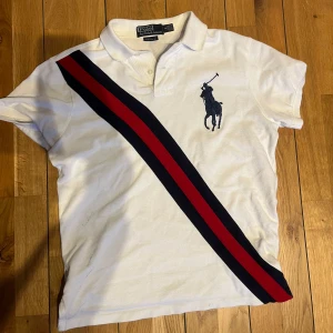  pikétröja från Ralph Lauren - Snygg vit pikétröja från Ralph Lauren med korta ärmar och klassisk krage. Tröjan har ett stort blått broderat logotypmotiv och diagonala ränder i rött och blått. Perfekt för en stilren look! Stolen S