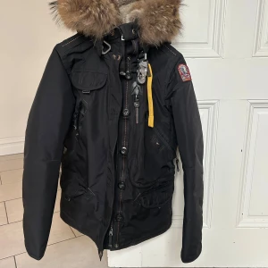 Svart jacka med pälsdetaljer från Parajumpers - I nyskick! Snygg svart jacka från Parajumpers med pälsdetaljer på huvan och flera praktiska fickor. Jackan har en dragkedja och knappar framtill samt en gul remdetalj. Perfekt för kyliga dagar. 