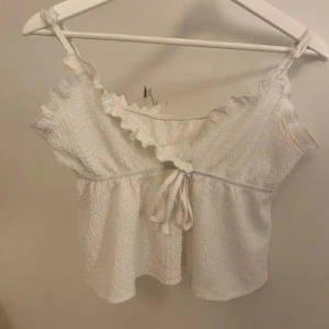 Vit topp med volang från Shein - Säljer en söt vit topp från Shein med volangdetaljer och knyt framtill. Toppen har smala axelband och en lätt textur i tyget. Perfekt för varma dagar!