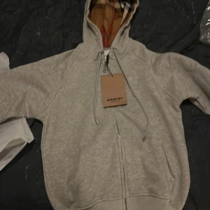 Grå huvtröja från Burberry - Säljer en stilren grå huvtröja från Burberry med dragkedja och klassiskt rutigt mönster i huvan. Tröjan har en broderad logga på bröstet. Helt oanvänd med lapparna kvar på, Priset är inte hugget i sten