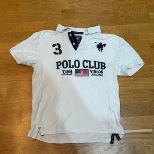 Vit kortärmad pikétröja från Polo Club - Snygg vit pikétröja från Polo Club med broderad polospelare på bröstet. Tröjan har tryck med texten 'Polo Club' och en amerikansk flagga. Perfekt för en sportig look!