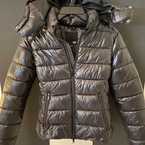 Svart pufferjacka med huva - Snygg svart pufferjacka med avtagbar huva och dragkedja framtill. Jackan har en glansig yta och två praktiska fickor med dragkedja. Jackan är sparsamt använd och har inga tydliga täcken på användning finns dock någon fläck vid kragen av smink 