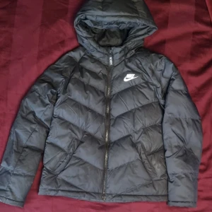 Svart dunjacka från Nike - Säljer en svart dunjacka från Nike med huva och dragkedja. Jackan har ett stilrent märke på bröstet och är perfekt för kyligare dagar. Den är långärmad och har en klassisk pufferdesign. Junior stl L 147-158cm