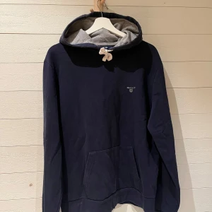 Mörkblå hoodie från Gant - Säljer en stilren mörkblå hoodie från Gant med grå insida i huvan och en praktisk magficka. Perfekt för en avslappnad look. Huvan har snörning för justerbar passform. Skick 8/10. Storlek L.