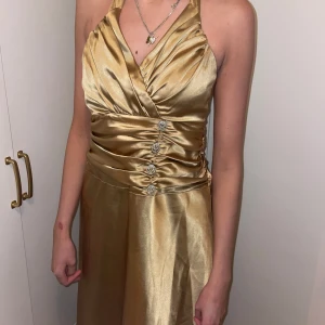 Guldfärgad klänning med halterneck - Elegant guldfärgad klänning med halterneck och omlottdesign. Klänningen har dekorativa knappar framtill och en öppen rygg med knytband. Perfekt för festliga tillfällen.