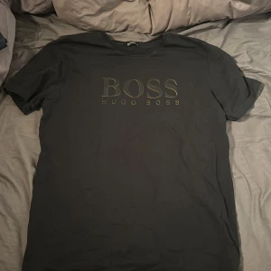Svart t-shirt från Hugo Boss pris kan diskuteras - Snygg svart t-shirt från Hugo Boss med deras ikoniska logga tryckt på framsidan. Perfekt för en stilren och avslappnad look. Kortärmad och tillverkad i mjukt material för extra komfort.