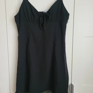 Svart klänning från H&M Divided - Snygg svart klänning från H&M Divided med knytning framtill. Klänningen har en enkel och stilren design med smala axelband och en lätt utställd kjol. Perfekt för olika tillfällen!