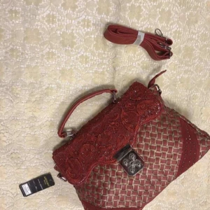 Röd handväska med spetsdetaljer - Snygg röd handväska från Arcade USA med vackra spetsdetaljer och flätat mönster. Väskan har ett handtag och en avtagbar axelrem. Perfekt för att ge en elegant touch till din outfit.  Stlk 38.00*26.00*10.50 cm , väska har bra utrymme, två huvud fickor samt 3 övriga fickor, en huvud blixtlås fack 