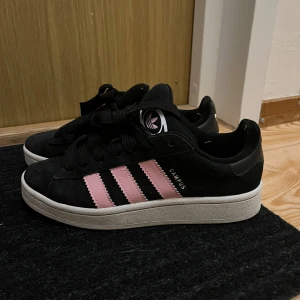 Svarta Adidas Campus med rosa ränder - Snygga svarta Adidas Campus sneakers med rosa ränder och vit sula. Fortfarande i bra skick och användes några gånger i våras